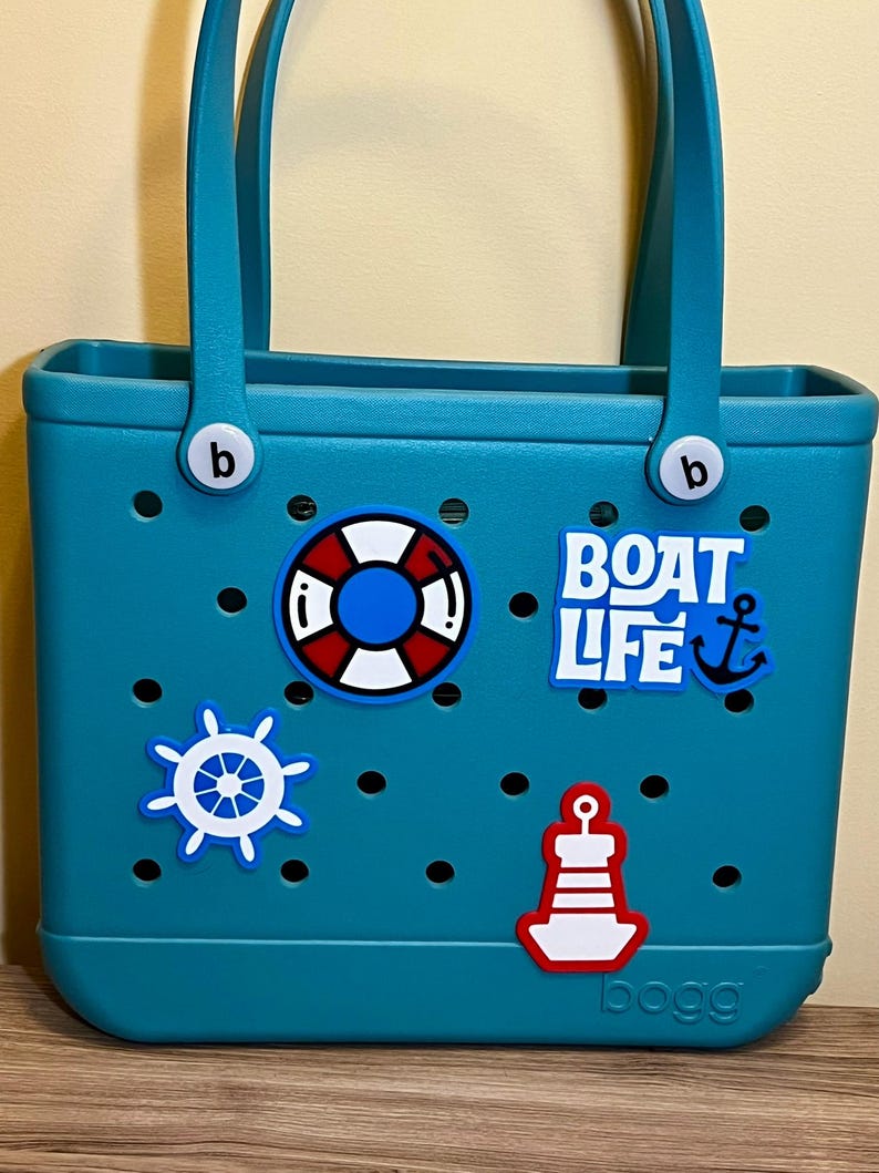 以下が含まれることがあります： 白と赤の航海をテーマにしたデカールが付いた、ティール色のトートバッグ。バッグにはハンドルがあり、側面に文字「b」があります。デカールには、救命浮き輪、船の舵、灯台、「BOAT LIFE」の文字と錨が含まれています。