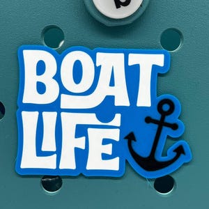 以下が含まれることがあります： 黒い錨のデザインの青と白の「BOAT LIFE」チャーム。チャームは、黒い「b」が付いた白いボタンが付いたティール色の背景に設定されています。この航海をテーマにしたチャームは、バッグやアクセサリーに最適です。