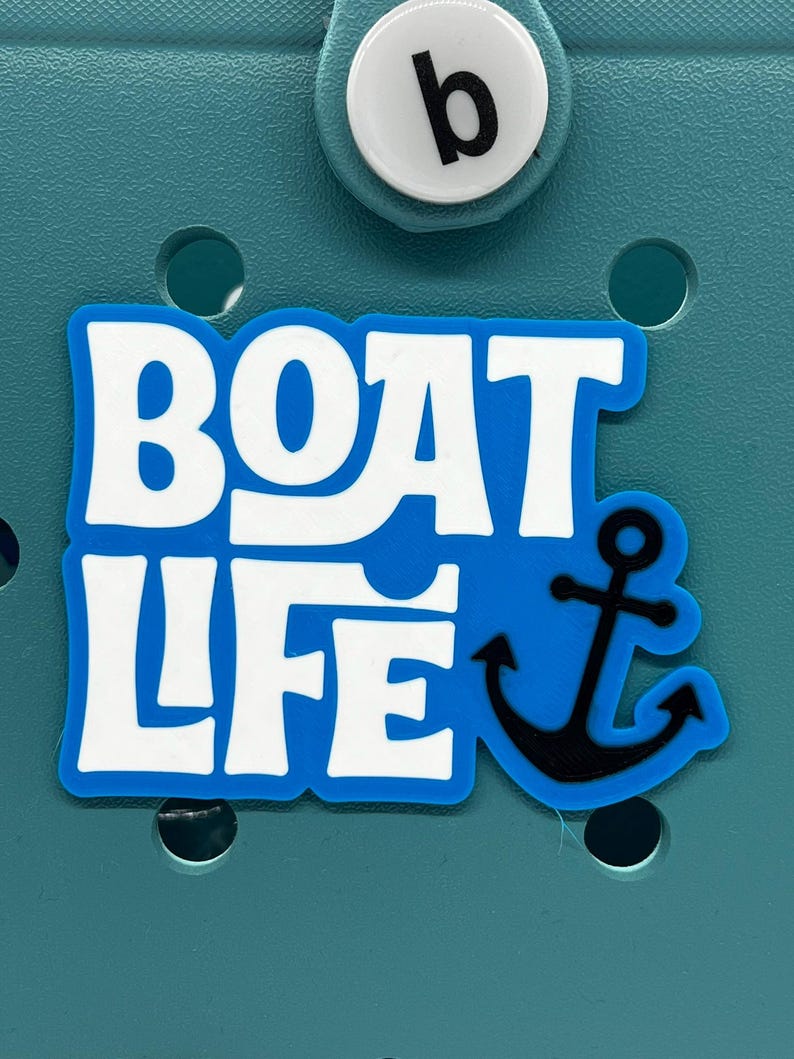 以下が含まれることがあります： 黒い錨のデザインの青と白の「BOAT LIFE」チャーム。チャームは、黒い「b」が付いた白いボタンが付いたティール色の背景に設定されています。この航海をテーマにしたチャームは、バッグやアクセサリーに最適です。