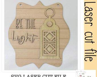 May include: A light-coloured wooden plaque with a scalloped edge featuring a laser-cut lantern design and the text "BE THE LIGHT" in raised lettering. The plaque includes two small holes at the top for hanging and a horizontal wood grain pattern.