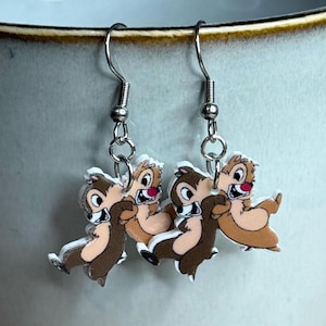 Puede incluir: Un par de pendientes de plata con un diseño de dibujos animados de Chip y Dale de Chip 'n Dale: Los rescatadores de Disney. Los pendientes están hechos de metal y tienen un acabado plateado.