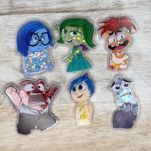 May include: Six colorful character stickers featuring characters from the animated film Inside Out. The characters are Joy, Sadness, Anger, Disgust, Fear, and Bing Bong. The stickers are made of clear plastic with glitter embedded inside.