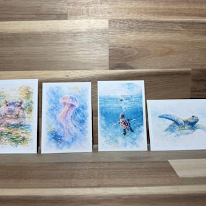 May include: Four watercolor art prints featuring aquatic animals. The prints depict a hippo in a pond, a jellyfish, a sea turtle swimming underwater, and a sea turtle. The prints are on white rectangular cards.