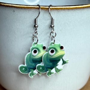 Puede incluir: Un par de pendientes colgantes con un camaleón verde de ojos grandes. El camaleón está impreso sobre un fondo blanco y los pendientes tienen ganchos plateados.