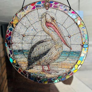 May include: A round stained glass suncatcher with a colourful mosaic border and a white pelican with a pink beak standing on a beach scene.