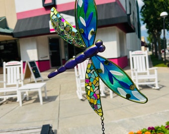 May include: A decorative dragonfly wind chime with a purple body and iridescent wings. The wings feature blue, green, and mosaic-style glass accents. A metal chain and spring connect the dragonfly to a hanging loop. The background includes flowers and buildings.