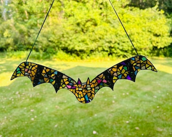 Op de afbeelding: Een vleermuis van glas-in-lood hangend aan een touwtje. De vleermuis is gemaakt van gekleurde glasstukken, waaronder geel, oranje, rood, groen, blauw en paars. De vleermuis is omrand met zwart.