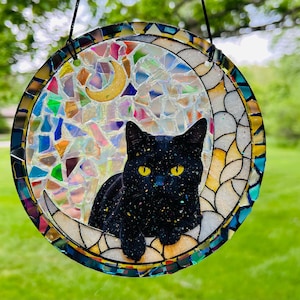 May include: A round, stained glass suncatcher featuring a black cat resting on a crescent moon. The cat has yellow eyes. The background is a mosaic of colorful glass pieces. The suncatcher is hanging from a black string.