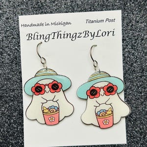 May include: A pair of ghost-shaped earrings. Each earring features a white ghost wearing a light blue hat and red flower-shaped sunglasses. The ghost is holding a pink bucket with flowers. The text "BlingThingzByLori" is visible.