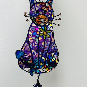 May include: A purple mosaic cat wind chime with blue and green accents. The cat is sitting with its tail curled around its legs. The wind chime has a series of blue glass beads hanging from it.