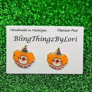 Puede incluir: Pendientes con forma de payaso con pelo naranja, nariz roja y gorro de fiesta. Los pendientes están sobre una tarjeta blanca con el texto "BlingThingzByLori" y "Handmade in Michigan". El fondo es una superficie verde texturizada.