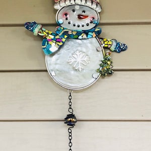 May include: A metal snowman wind chime with a mosaic design. The snowman is wearing a blue and green scarf and a colorful hat. The chime has a silver bell at the bottom.