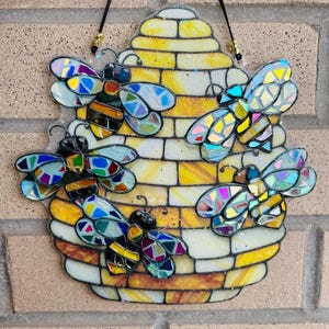 May include: A decorative stained glass piece featuring a yellow and white beehive with four colorful bees. The bees have iridescent wings and black and yellow striped bodies. The piece is designed for hanging.