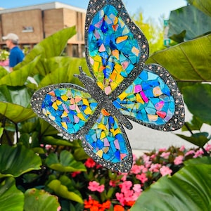 May include: A blue and yellow mosaic butterfly with glitter accents hanging from a string. The butterfly is in front of a green leafy plant and red flowers.