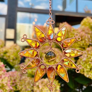 May include: A copper suncatcher with a glass orb in the center. The suncatcher is made of stained glass pieces in shades of yellow, orange, and red. The suncatcher is hanging from a chain.
