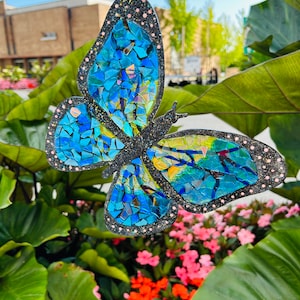May include: A blue mosaic butterfly with glitter accents hanging from a string in front of a green leafy plant.