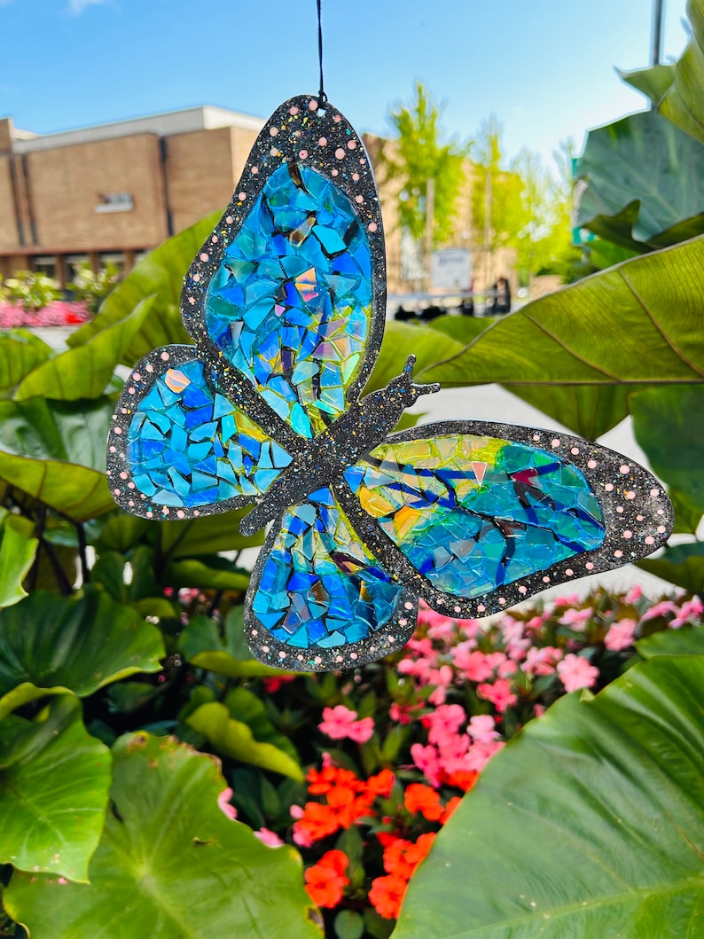 May include: A blue mosaic butterfly with glitter accents hanging from a string in front of a green leafy plant.