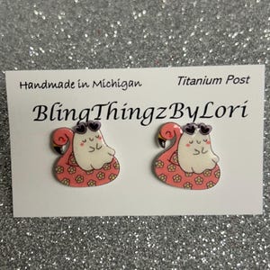 May include: A pair of flamingo-shaped earrings with sunglasses, set on a white card. The earrings have a pink body with a floral pattern and a white head. The card reads "Handmade in Michigan" and "BlingThingzByLori."