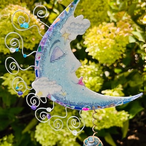 May include: A blue crescent moon-shaped wind chime with a whimsical face, clouds, and stars. The chime has a silver wire with purple and blue beads and a blue glass orb hanging from the bottom. The text "Sweet dreams delivered on moonbeams" is written on the moon.