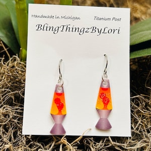 May include: Lava lamp earrings with orange and yellow liquid and red heart-shaped accents. The earrings have a purple base and are suspended from silver hooks. The card behind the earrings reads "BlingThingzByLori."
