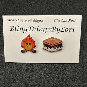 以下が含まれることがあります： 手作りのチタンポストピアス。キャンプファイヤーとスモアのデザイン。キャンプファイヤーはオレンジと黄色の炎で、笑顔の顔があります。スモアはグラハムクラッカー、チョコレート、マシュマロ、笑顔の顔があります。