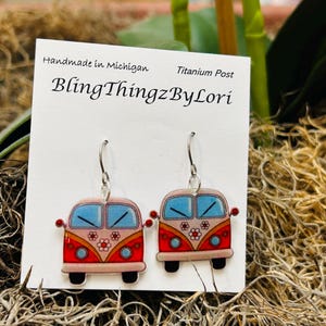 May include: A pair of earrings featuring a red, orange, and pink camper van design with blue windows and flower accents. The earrings are on a white card with the text "BlingThingzByLori" and "Handmade in Michigan".