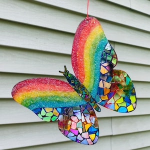 May include: A rainbow-colored butterfly made of mosaic glass tiles. The butterfly is hanging from a red string and has a glittery finish.