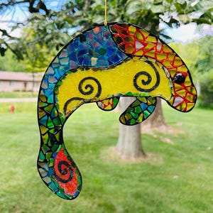 May include: A dolphin-shaped suncatcher crafted from colorful mosaic glass pieces. The body features a central yellow section with black spiral details, surrounded by blue, green, and red glass fragments. A single black glass eye is visible.