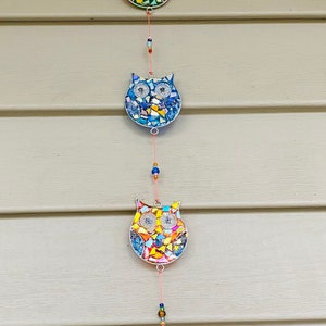 May include: A hanging owl decoration with five mosaic owl figures in various colors. Each owl has a silver outline and is strung on a red cord with small beads. The bottom has a red, oval-shaped glass piece.