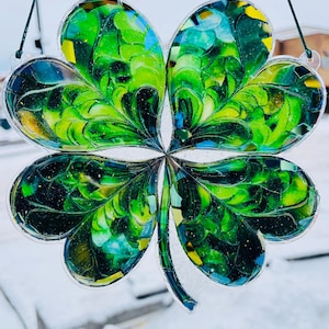 Puede incluir: Un trébol de cuatro hojas de vidriera con tonos verdes, azules y amarillos. El trébol está suspendido por una cuerda verde. El vidrio tiene un aspecto de mosaico, con patrones y texturas intrincados.