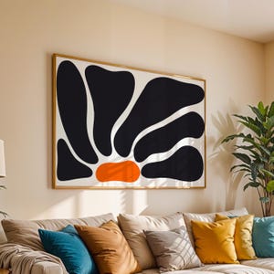 Op de afbeelding: Groot ingelijst abstract kunstwerk met een gedurfd zwart-wit bloemmotief met een oranje centrum. Het kunstwerk wordt weergegeven boven een bank met kleurrijke sierkussens, wat een moderne en stijlvolle woonkameresthetiek creëert.