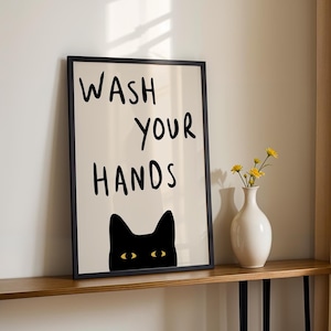 Puede incluir: Un cuadro enmarcado con las palabras "WASH YOUR HANDS" en negro, sobre una silueta de gato negro con ojos amarillos. El cuadro está en una estantería de madera, con un jarrón blanco con flores amarillas.