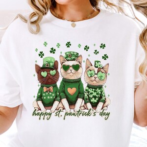 May include: White t-shirt with a graphic of three cats wearing green sweaters, hats, and sunglasses. The cats are sitting together and the text "happy st. pawtrick's day" is below them.