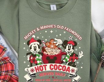 May include: Green sweatshirt with a Christmas-themed design featuring Mickey and Minnie Mouse. The text reads "Mickey & Minnie's Old Fashioned *HOT COCOA* *SERVED DAILY* Warm Up Here - Open All Winter".