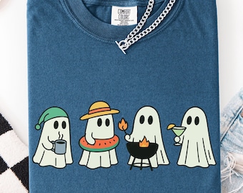 May include: A blue Comfort Colors t-shirt featuring a graphic of four cartoon ghosts. Each ghost is dressed for a different activity: drinking coffee, wearing a floatie, grilling, and holding a cocktail. A silver chain necklace is draped over the shirt.