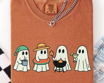 May include: A rust-colored Comfort Colors t-shirt featuring four cartoon ghosts, each with a unique accessory: a beanie with a mug, a sun hat with a watermelon float, a flame with a grill, and a cocktail. A silver chain necklace rests on the shirt.