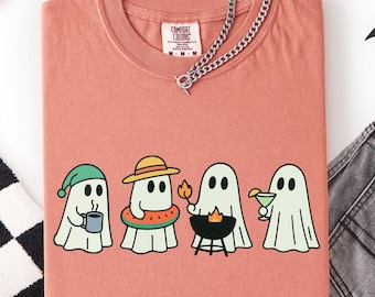 May include: A coral-colored Comfort Colors t-shirt featuring a graphic of four cartoon ghosts. Each ghost is depicted with a different accessory: a coffee mug, a hat and inner tube, a grill, and a cocktail. A silver chain is draped over the shirt.