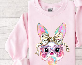May include: A pink sweatshirt with a colorful cartoon bunny wearing a bow and sunglasses blowing a bubble. The bunny has rainbow colored fur and a white face.