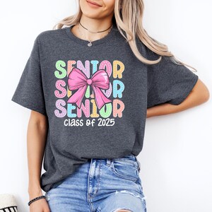 May include: A dark grey t-shirt with a colourful graphic design that says "Senior" three times with a pink bow in the middle. The text "Class of 2025" is printed below the graphic.