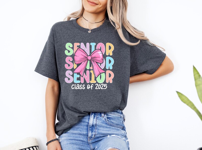 May include: A dark grey t-shirt with a colourful graphic design that says "Senior" three times with a pink bow in the middle. The text "Class of 2025" is printed below the graphic.