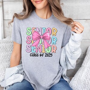 May include: A grey t-shirt with a colourful graphic design that says "Senior" with a pink bow in the middle and "Class of 2025" at the bottom.