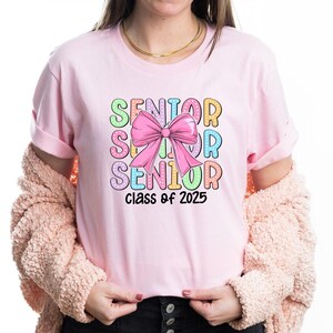 May include: Pink t-shirt with a colourful graphic that says "Senior" with a pink bow and "Class of 2025" underneath.