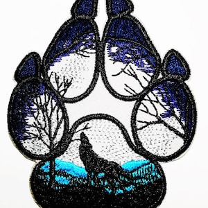 Può includere: Toppa ricamata a forma di impronta di zampa di lupo. L'impronta è nera con dettagli blu e bianchi. Il disegno presenta un lupo che ulula, alberi e un paesaggio montano.