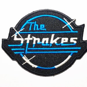 May include: Black and blue embroidered patch with the text "The Strokes" in a white outline. The patch has a white X design with a blue circle around it.