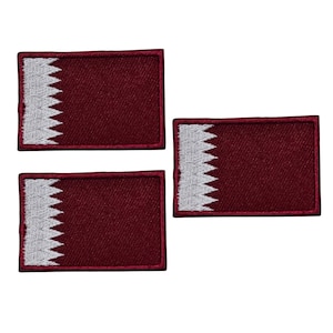 Puede incluir: Tres parches rectangulares con la bandera de Qatar. Cada parche tiene un fondo granate con un borde blanco dentado en el lado izquierdo. Los parches están dispuestos sobre un fondo blanco.