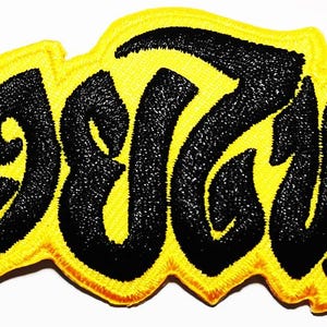 May include: Embroidered patch featuring black text on a yellow background. The text is in a stylized font, with a wavy yellow border. The patch is designed to be sewn or ironed onto clothing or accessories.
