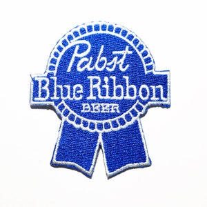 May include: A blue and white embroidered patch featuring the Pabst Blue Ribbon beer logo. The patch is in the shape of a ribbon with the words "Pabst Blue Ribbon" and "Beer" embroidered in white.