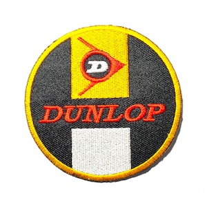 May include: A round black and yellow embroidered patch with the word "DUNLOP" in red lettering. A stylized yellow and red "D" is above the word.