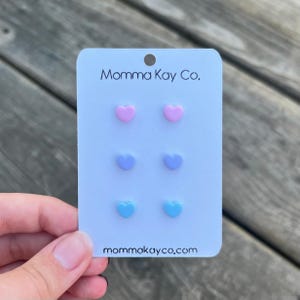 May include: A white display card labeled Momma Kay Co. at the top and mommakayco.com at the bottom, featuring three pairs of small heart-shaped stud earrings in pastel pink, light purple, and light blue arranged in a vertical column.