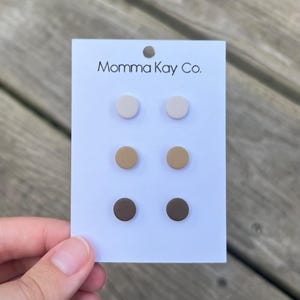 May include: A set of six small, round stud earrings in three colors: white, beige, and brown. The earrings are displayed on a white card with the text "Momma Kay Co."